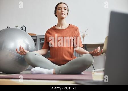 Pregnant woman sitting on the floor in front of laptop watching online lesson and meditating in the room Stock Photo