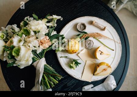 Wedding ring lies in a box on a white plate next to pears and green twigs. A plate with a bouquet of flowers tied with a ribbon stands on a wooden Stock Photo