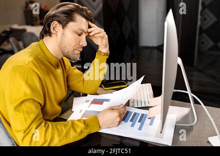 Serious male freelancer in yellow shirt doing some paperwork, learning financial graphs for important work report. Concentrated businessman is sitting Stock Photo
