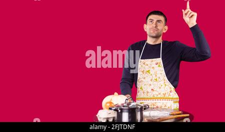 Banner, long format over red background. Confident young man showing forefinger up at kitchen table, home. cook baker man in apron. Correct new idea Stock Photo