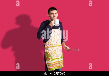 Thoughtful young man wearing kitchen apron looking to camera while holding white blank mock up cup of tea in kitchen and a cooking utensil tools Stock Photo