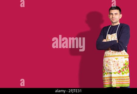 Banner, red background. Caucasian brunette young handsome chef man, in colorful apron crossed arms looking confident at the camera. Plenty space for Stock Photo