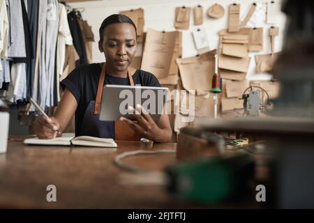 African female leather worker writing notes and using a tablet Stock Photo