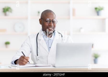 Professional help concept. Friendly black male doctor in white unifrom sitting at table with laptop at his office Stock Photo