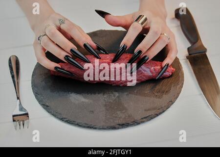 Female hands with very long black nails touching uncooked steak. Stock Photo