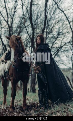 Woman in chain mail in image of medieval warrior holds her horse by the reins among forest. Stock Photo