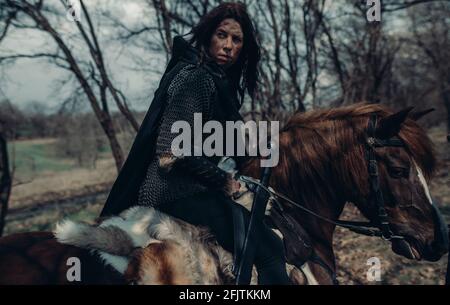Woman in chain mail in image of medieval warrior sits horseback among forest. Stock Photo