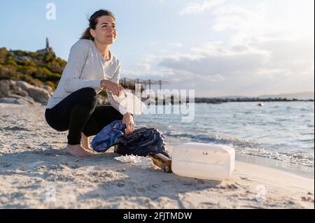 Smiling young woman on the beach recollecting plastic waste and put into a bag. Stock Photo