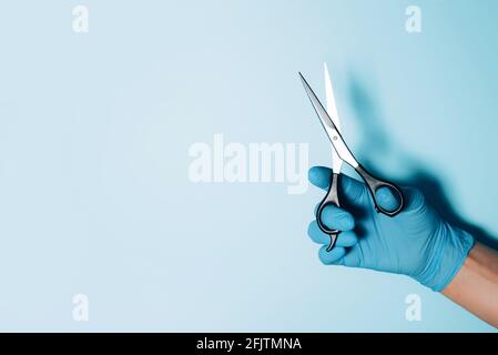 Hairdresser hand in medical glove holding scissors on blue background. New normal concept. Copy space. Stay safe. Health protection equipment during Stock Photo