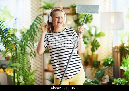 smiling modern 40 years old woman in striped shirt with mop and headphones doing household work in the modern living room in sunny day. Stock Photo