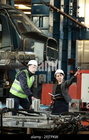 Portrait of a female factory manager in a white hard hat and business suit and factory engineer in work clothes. Controlling the work process at the airplane manufacturer. Stock Photo