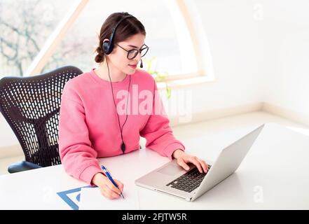 Young businesswoman in headset speaking by conference call while sitting behind her laptop. Home office. Stock Photo