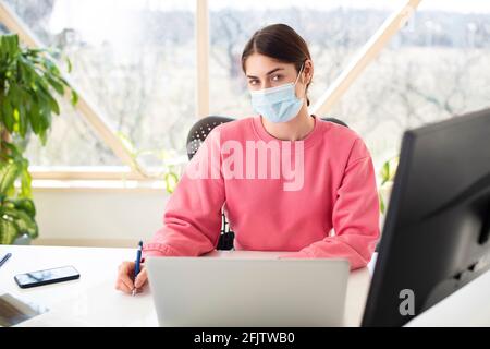 Shot of beautiful woman wearing face mask while sitting behind her computer and working. Home office. Stock Photo
