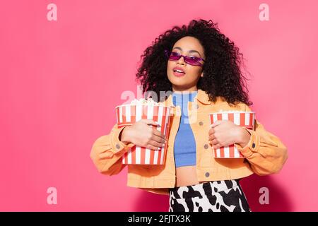 african american woman in sunglasses holding buckets with popcorn on pink Stock Photo