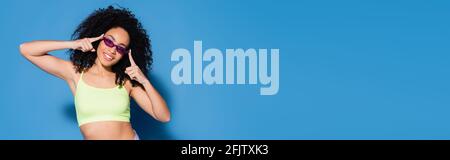 happy african american woman adjusting sunglasses with fingers on blue, banner Stock Photo
