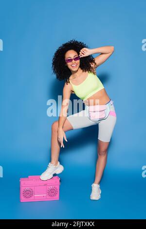 full length of cheerful african american woman in sunglasses and waist bag posing near pink boombox on blue Stock Photo