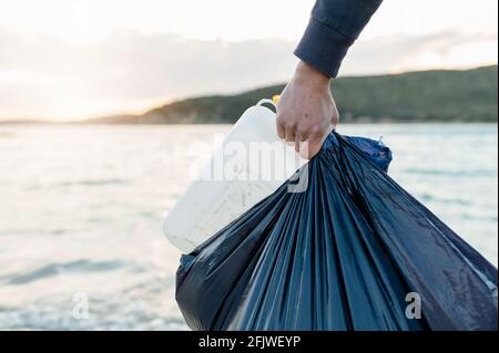 Unrecognizable man arm holding a bag full of garbage and a plastic bottle with sea on background. Volunteer work and cleaning beach from pollution con Stock Photo