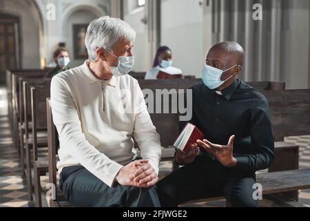 African young priest in mask talking to senior man while they sitting on bench in the church Stock Photo