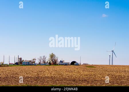 Power windmills spin under an almost cloudless sky and over early spring farm fields and nearby farm buildings. Stock Photo