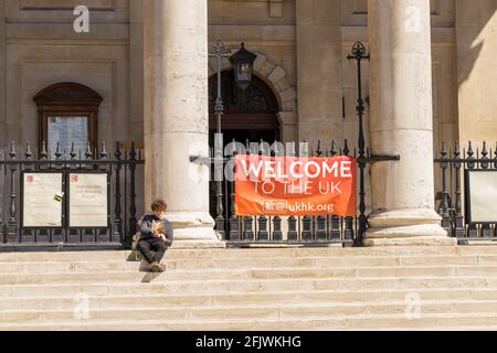 Man sitting in the sun reading a book in front of a Welcome To The UK sign on the steps of St Martin in The Fields Church on Charing Cross Road London Stock Photo