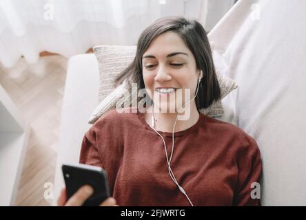 Smiling woman is lying on the sofa and having a video call with her black smartphone and her white headphones in her living room at home Stock Photo