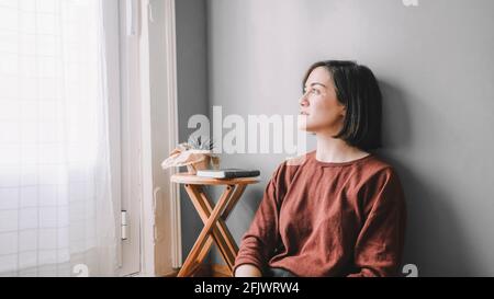 Smiling woman sitting on the wooden floor looks at the window with white curtains and a wooden bench with a green notebook and a cactus Stock Photo
