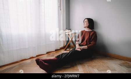 Smiling woman sitting on the wooden floor of her room looking at the window with white curtains and a wooden bench with a green notebook and a cactus Stock Photo