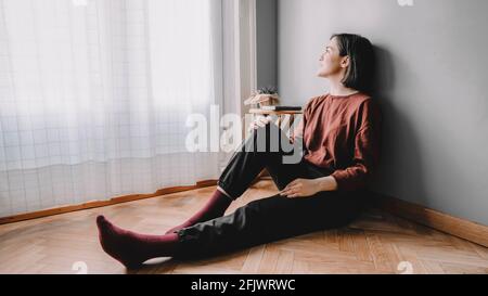 Smiling woman sitting on the wooden floor of her room looks at the window with white curtains and a wooden bench with a green notebook and a cactus at Stock Photo