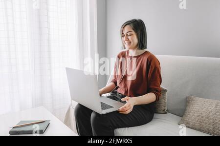 Woman with headphones connected to a laptop is smiling and looking to it sitting on the sofa near the white curtains window at home Stock Photo