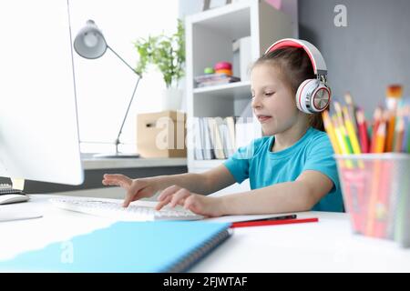 Little girl in headphones in front of computer is typing on keyboard Stock Photo