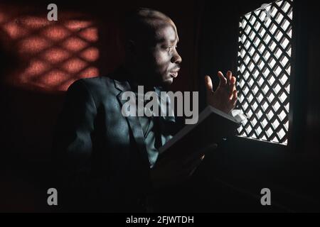 African young priest reading the Bible while sitting in confessional Stock Photo