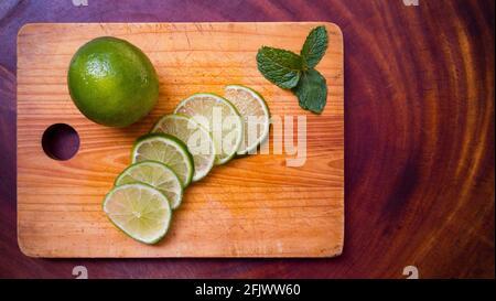 Green lemon, slices of lemon and mint on cutting board and wooden background. Different shades of wood. Top view. Stock Photo