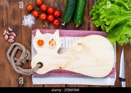 Cutting wooden board in the form of a cat with eyes made from fresh cherry tomatoes on a wooden table. Handmade chopping board. Fresh tomatoes, cucumb Stock Photo