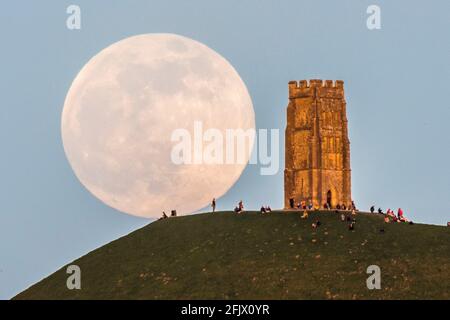 Glastonbury, Somerset, UK.  26th April 2021.  UK Weather.  The full pink super moon rises up from behind Glastonbury Tor in Somerset on a clear evening as people sit on top and watch it rise.  Picture Credit: Graham Hunt/Alamy Live News Stock Photo