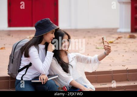 Two students with beaming smiles are posing for selfie shot, Asian attractive lady is taking in university or campus. Stock Photo