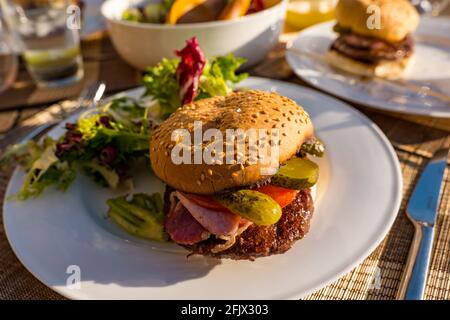 Beef burger with bacon, tomato & dill pickle on a white plate with salad on an outdoor table in sunshine Stock Photo