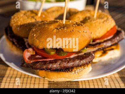 Close up of beef burgers with bacon, tomato & dill pickle on a white plate outdoors in sunshine Stock Photo
