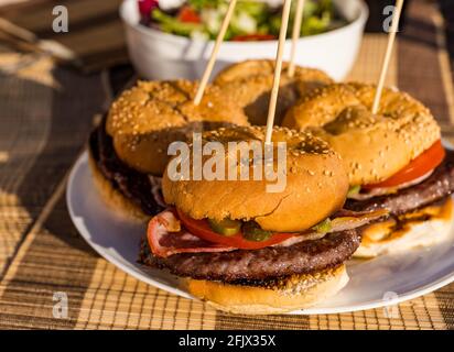 Beef burgers with bacon, tomato & dill pickle on plate with salad bowl outdoors in sunshine Stock Photo
