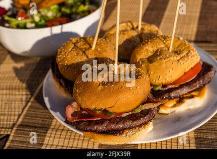 Beef burgers with bacon, tomato & dill pickle on plate with salad bowl outdoors in sunshine Stock Photo