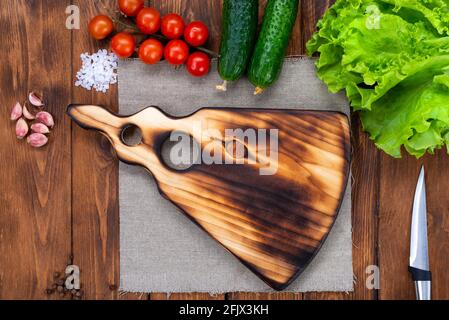 Cutting wooden board on a wooden table. Handmade chopping board. Fresh tomatoes, cucumbers and a cutting board. View from above. Stock Photo