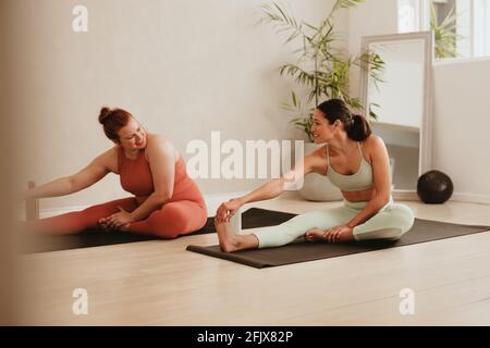 Two females exercising together in fitness studio. Women in sports clothing doing leg stretching workout at gym. Stock Photo