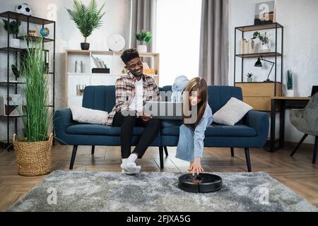 Multicultural young couple using modern laptop and robot vacuum cleaner for doing household. African man and caucasian woman relaxing together on soft couch. Stock Photo
