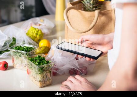 Online home food delivery of fresh vegetables and fruits. Young man holding phone and checking order list. Reusable bag with bio vegetables on white Stock Photo