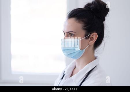 Thoughtful female doctor standing with hands in pockets while looking through window at hospital corridor Stock Photo