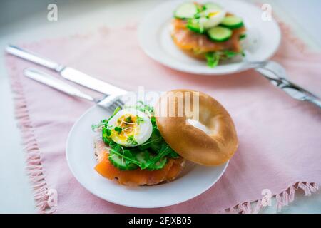 Fresh bagel sandwiches with salmon, soft egg, salad, microgreen sprouts, cucumber and cream cheese served on pink napkin with cutlery on the white Stock Photo