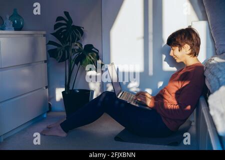 Mixed race woman sitting on the floor in her bedroom using a laptop in sunset light and shadows. Remote work at home. Woman scrolling social networks Stock Photo