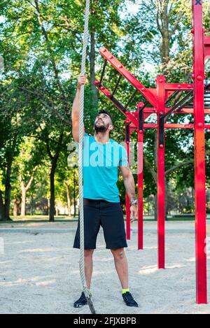 Dark-haired athlete with beard ready to climb a rope. Park background and calisthenics bars. Stock Photo