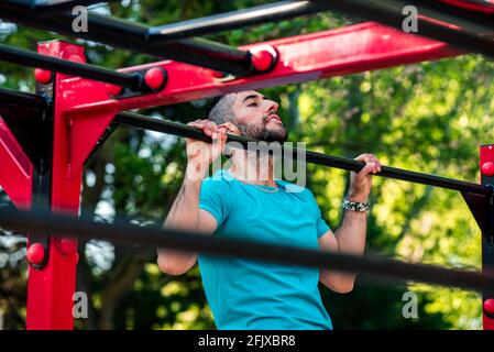 Dark-haired athlete with beard doing a pull-up on a calisthenics bar. View between bars. Outdoor crossfit concept. Stock Photo