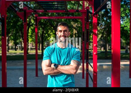 Portrait of a brunette bearded athlete with crossed arms. Red calisthenics bars in the background . Outdoor fitness concept. Stock Photo
