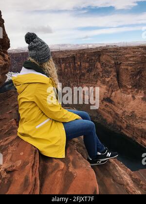 Woman sitting on the rocks of the Grand Canyon National Park in Arizona. Woman wearing a yellow rain jacket and black new balance sneakers. Stock Photo